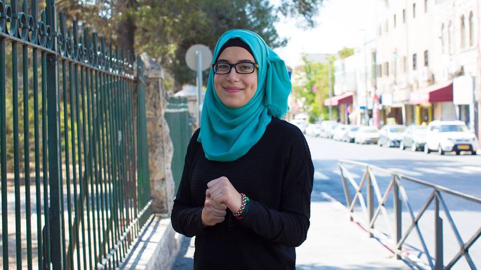 A woman dressed in a black sweater and a green hijab, wearing dark-rimmed glasses