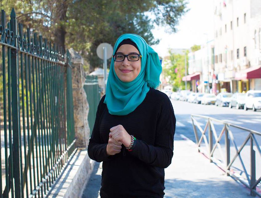 A woman dressed in a black sweater and a green hijab, wearing dark-rimmed glasses