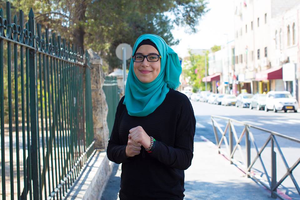 A woman dressed in a black sweater and a green hijab, wearing dark-rimmed glasses