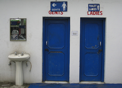 Two blue doors against a white wall, one having 'gents' and the othe rhaving 'ladies' written over it. beside them, there is a white sink.