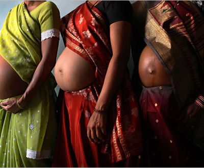 The pregnant bellies of three women standing in a line. One of them wears a green saree, the other two wear red sarees.