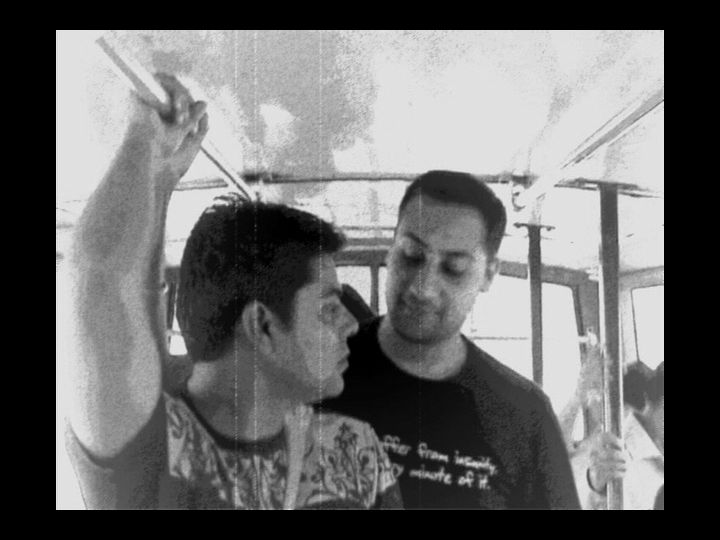 Two men in a public bus facing each other, one of them standing behind the other and they are looking at each other fondly.