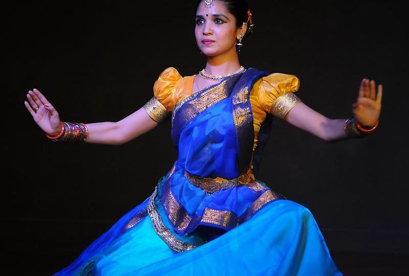 A woman wearing a blue saree and an orange blouse performing Kathak. She is wearing colourful bangles and jewellery on top of her forhead, ears, and neck. She is wearing kajal, a black bindi, and her hair is tied.