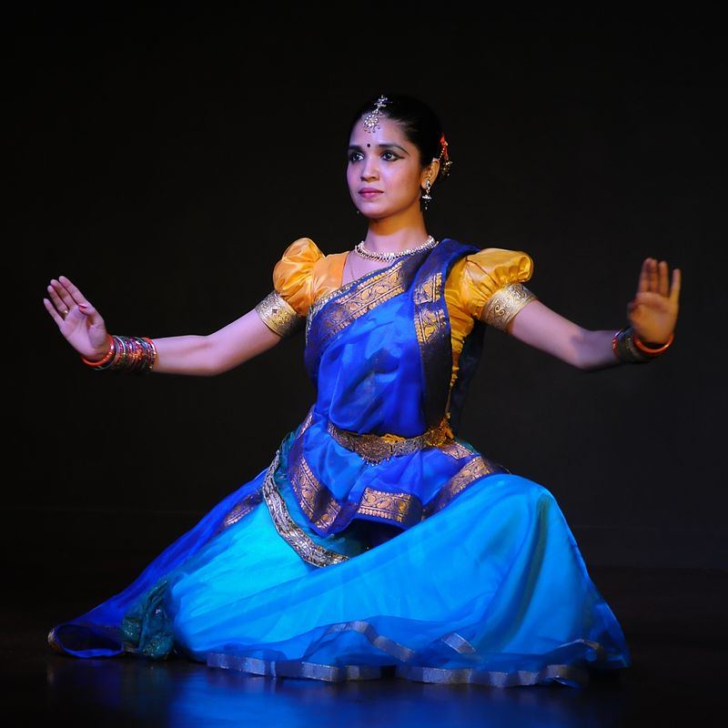 A woman wearing a blue saree and an orange blouse performing Kathak. She is wearing colourful bangles and jewellery on top of her forhead, ears, and neck. She is wearing kajal, a black bindi, and her hair is tied.