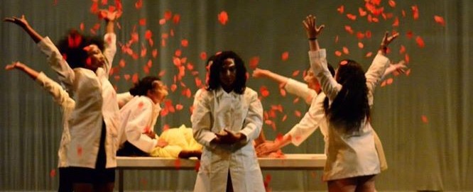 Stage performance of a few women in white coats holding down a woman on a table to commit Female Genital Mutilation, while others throw red rose pertals in air. One woman stands in the front and centre of the stage looking solemnly.