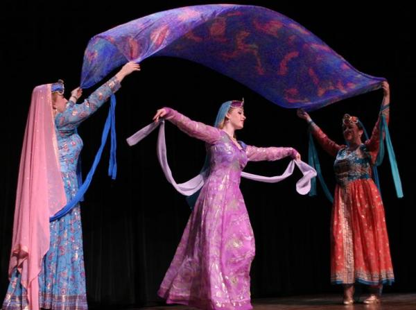 Stage performance of Persian dance. Two women hold a chunni above the head of a woman dancing in between them. They all wear colourful full-length, full sleeves clothing.