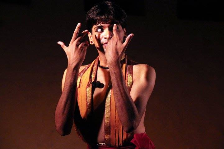 A boy performing Bharatnatyam on stage. He wears kajal, and an orange dupatta hung around his neck falls from both sides till his waist. The photo ends at his torso.