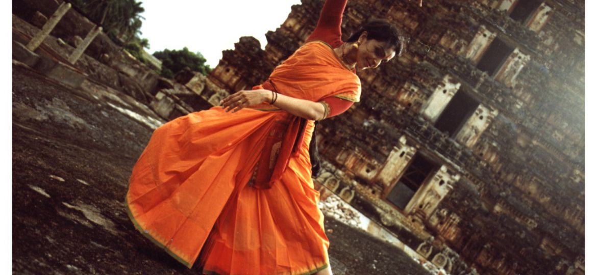 Anita Ratnam in an orange saree doing Bharatnayam outside an old temple.