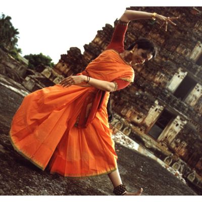 Anita Ratnam in an orange saree doing Bharatnayam outside an old temple.
