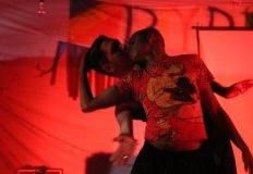 Two men dancing on a stage with red flourescent lights. The dance pose shows the man in front turning over his shoulder to kiss the man standing behind him.