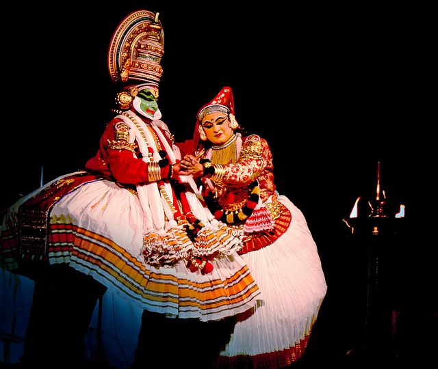 A man and a woman perform Kathakali on stage, wearing typical Kathakali costumes of red and white and elaborate headgear. A lamp is lit on the stage. Their makeup and dresses show beautifully in contrast to the darkened stage.