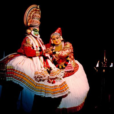 A man and a woman perform Kathakali on stage, wearing typical Kathakali costumes of red and white and elaborate headgear. A lamp is lit on the stage. Their makeup and dresses show beautifully in contrast to the darkened stage.