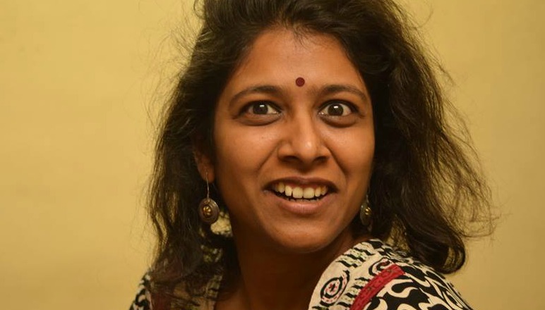 A woman's headshot. She is looking brightly at the camera. She is wearing a red bindi, earrings, and a black and white-coloured suit. Her hair is left open.