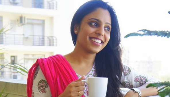 A young woman in a white Indian suit and red chunni standing on a balcony with a coffee mug in her hands. Her hair is let down and pulled over the left shoulder. She is smiling broadly.