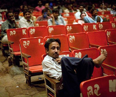 A man sits leisurely with his legs on his chair in a theatre. The audience chairs are coloured red, and are numbered. Many more men who are out-of-focus in this picture sit in similar fashion in rows behind him, looking at the screen.