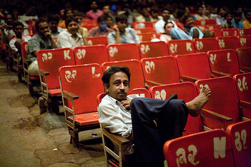 A man sits leisurely with his legs on his chair in a theatre. The audience chairs are coloured red, and are numbered. Many more men who are out-of-focus in this picture sit in similar fashion in rows behind him, looking at the screen.