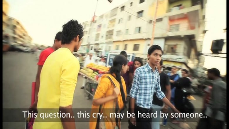 People moving on an Indian road. Fruitsellers, young boys, a woman. On both side are yellow-coloured buildings for flats. Subtitle reads, "This question... this cry... may be heard by someone."
