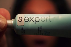 A woman holding a green-cloured tooothpaste tube, on which is written "expert" with the letter "s" prefixed before it manually by a pasting a piece of paper after drawing an S.