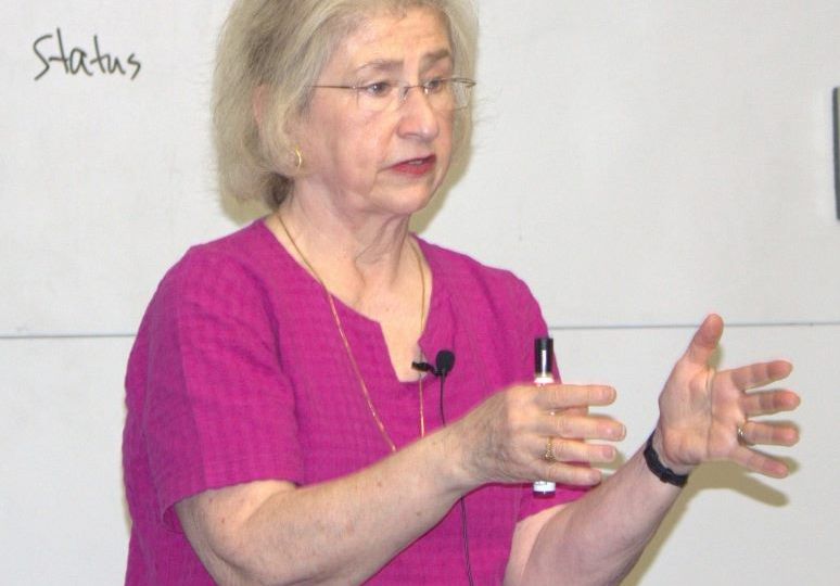 A picture of a caucasian woman with white hair wearing a magenta top and holding her hands up