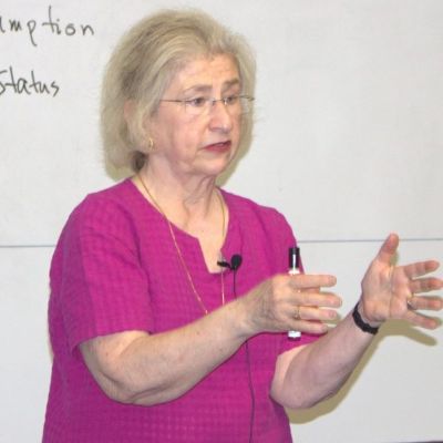 A picture of a caucasian woman with white hair wearing a magenta top and holding her hands up