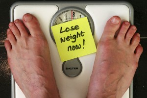 Photo of feet standing on a weighing machine. On the scale is a sticky note that reads in black cursive, "lose weight now!"