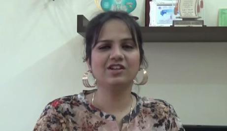 A photo of Nidhi Goyal with her hair tied back, wearing golden earrings, golden necklace, and a peach top. She is speaking.