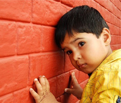 A male child dressed in a yellow shirt leaning against a red brick wall