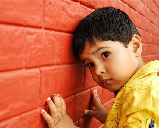 A male child dressed in a yellow shirt leaning against a red brick wall