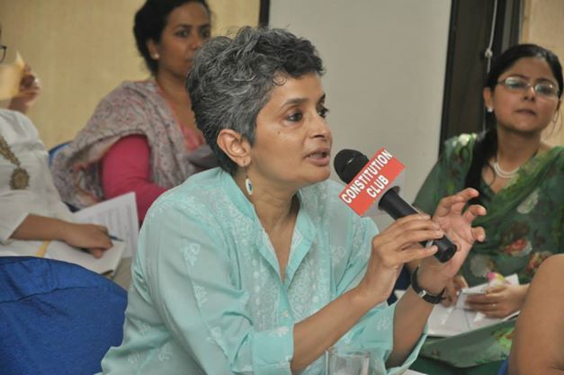 Nivedita Menon speaking into a mike of Constitution Club at a gathering. She is wearing a green kurta, and earring. She and others are sititng on ground cross-legged.