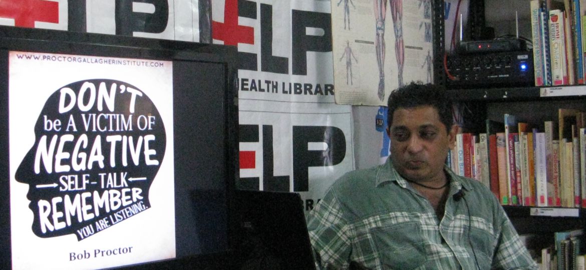 A man in a checkered shirt sits at a dek where a placard shows the rough outline of the human brain with messages about the importance seeking help for bad mental health written on it