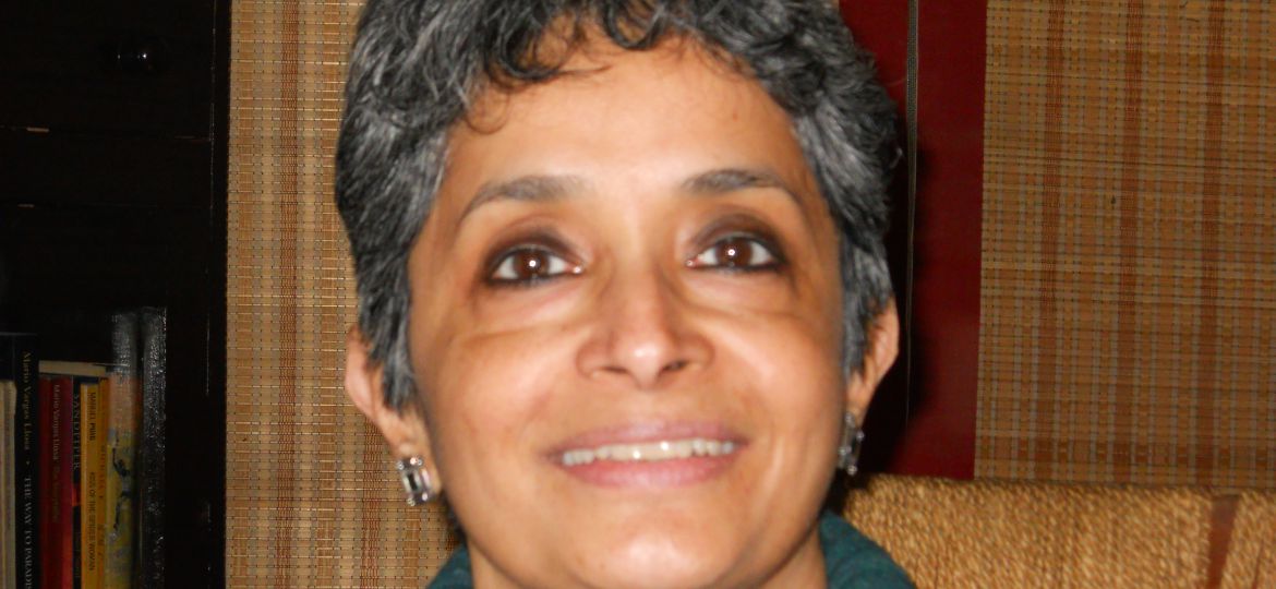 Nivedita Menon, wearing a green turtle-neck sweater, sitting on a chair, smiling.