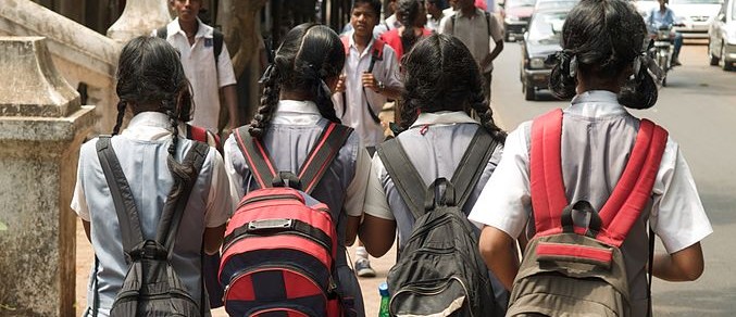 Children going to or returning from school. The focus is on four girls in grey and white school uniforms, wearing two braided plaits, and school bags on their shoulders, walking with their backs to us.