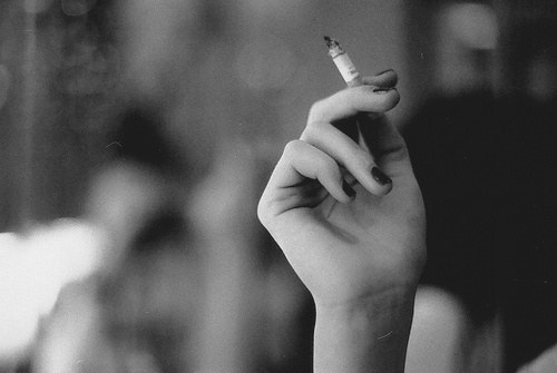 A black-and-white photo of a hand resting by an elbow, holding a cigarette. The nails are painted in a dark solid colour.