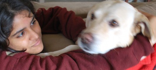 A girl hugging her dog. She is wearing maroon sweatshirt, and the white-coloured dog wears red warm piece of clothing.