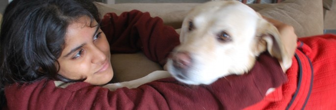 A girl hugging her dog. She is wearing maroon sweatshirt, and the white-coloured dog wears red warm piece of clothing.