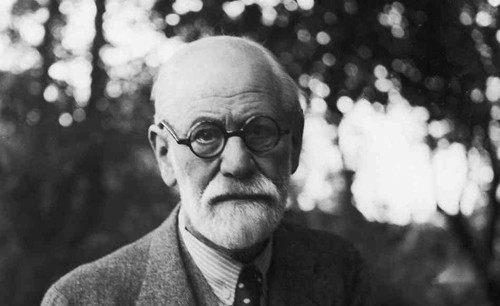 Black and white picture of psychologist Sigmund Freud. He has a white beard, is wearing dark round glasses along with a tweed jacket