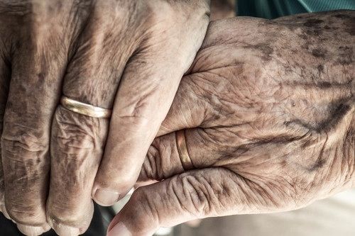A pair of hands with wrinkles on them holding each other. One of the hands is wearing a gold band on their ring finger