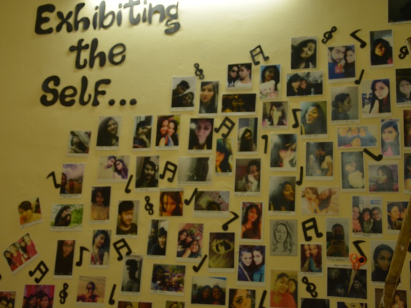 Cut-outs of selfies of various people stuck on a yellow wall. On the top is pasted in black cut-out paper, "Exhibiting the self". Cut-outs of music symbols are pasted in between the selfies.