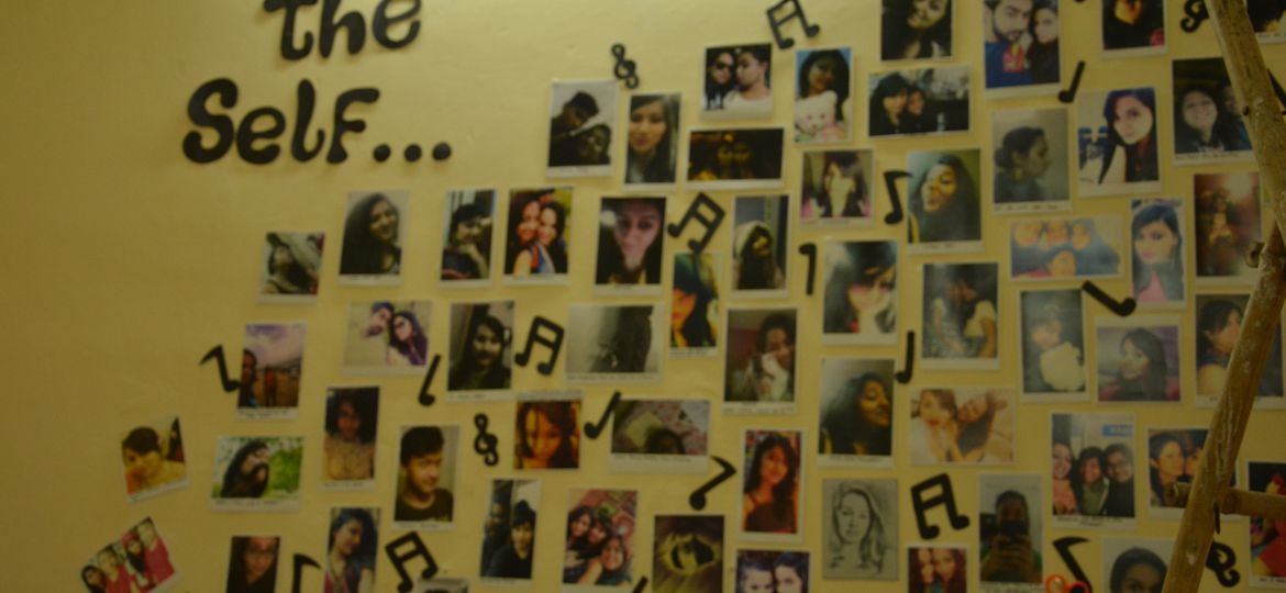 Cut-outs of selfies of various people stuck on a yellow wall. On the top is pasted in black cut-out paper, "Exhibiting the self". Cut-outs of music symbols are pasted in between the selfies.