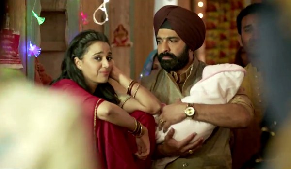 Still from film 'Tanu Weds Manu Returns'. A woman and a man in a wedding. The man holds a baby wrapped in a white cloth in his arms. The woman wears a red Indian suit, red bangles, and has open hair. The man wears a red turban and golden kurta.