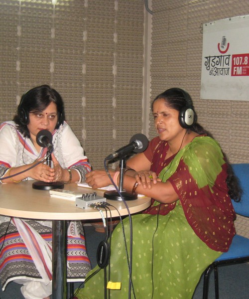 Two middle-aged women sitting in a recording room, wearing headphones, speaking into their table mikes. On the wall behind them is a poster that reads, "Gurgaon Awaaz 107.8 FM". One is wearing a green and maroon saree, and the other is wearing a white and pink Indian suit.