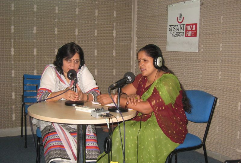 Two middle-aged women sitting in a recording room, wearing headphones, speaking into their table mikes. On the wall behind them is a poster that reads, "Gurgaon Awaaz 107.8 FM". One is wearing a green and maroon saree, and the other is wearing a white and pink Indian suit.