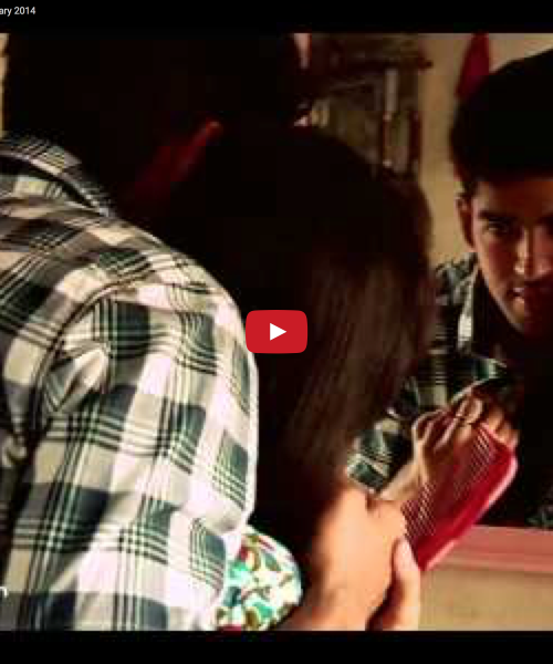 Still from a video. A boy hugs a woman from behind. They look at each other in the mirror in front of them. The woman is holding a comb in her hand. The boy is wearing a checked black and white shirt.