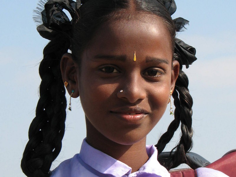 Photo of an Indian girl in a school uniform, carrying her schol bag on her shoulders. Her hair are neatly set in two braids, tied with a black ribbon.