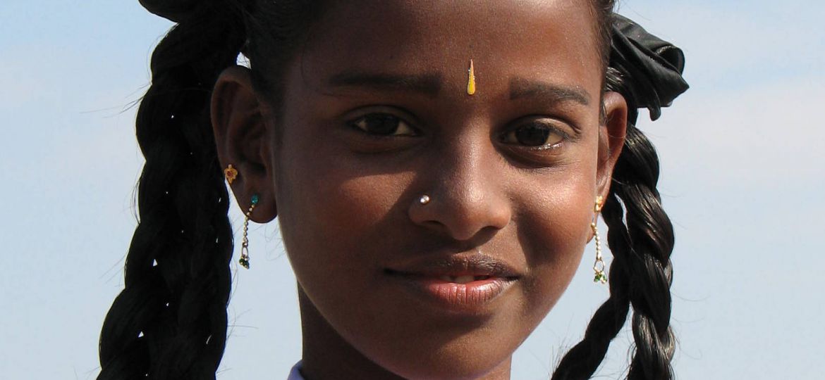 Photo of an Indian girl in a school uniform, carrying her schol bag on her shoulders. Her hair are neatly set in two braids, tied with a black ribbon.