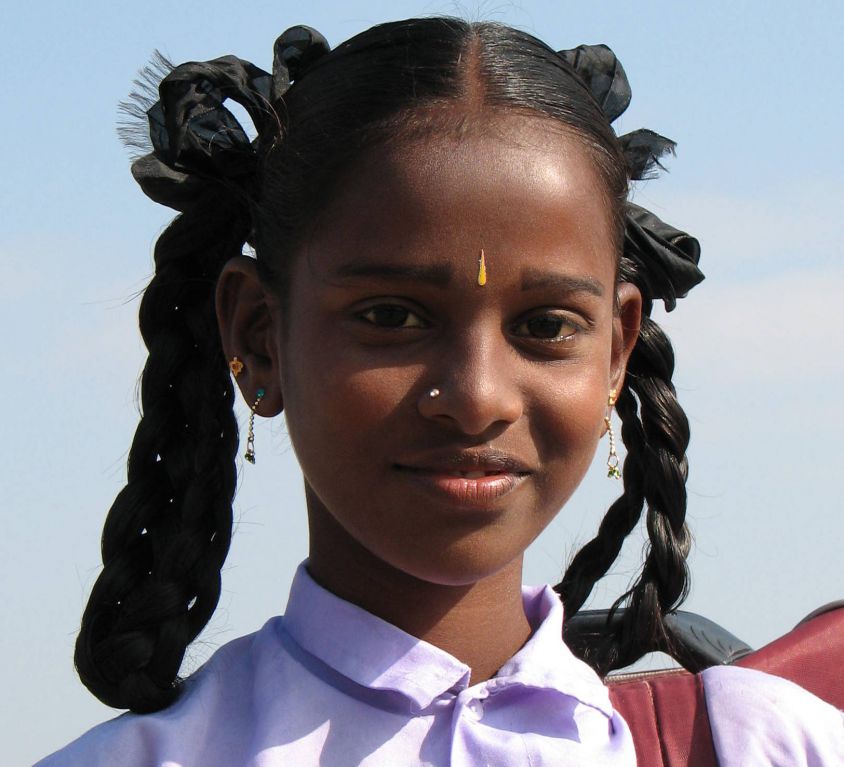 Photo of an Indian girl in a school uniform, carrying her schol bag on her shoulders. Her hair are neatly set in two braids, tied with a black ribbon.
