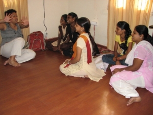 A woman sitting on floor, teaching something to some girls sitting in front of her on the floor in a house.