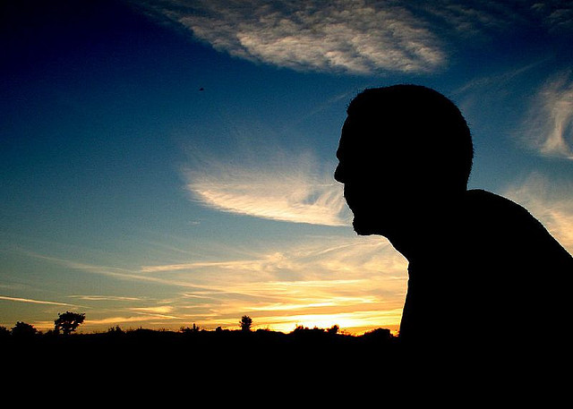 Silhouette of a man sitting in an open landscape. Sunset, windy weather, and cloudy sky could be seen behind him.