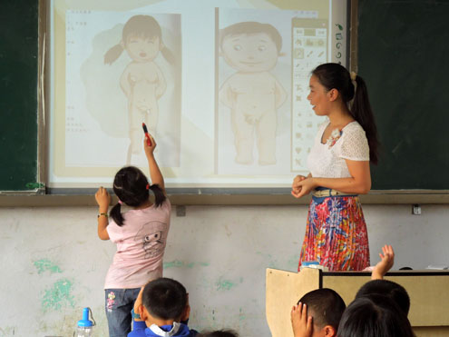 Photo of a class studying anatomy. A little girl pointing to the vagina of the person in the projected image on the board. The teacher stands on the side, looking on.