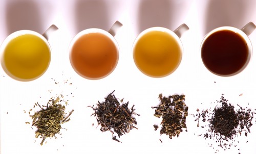 Four cups of different flavoured teas kept in a row horizontally. Next to each is kept corresponding dry tea leaves.